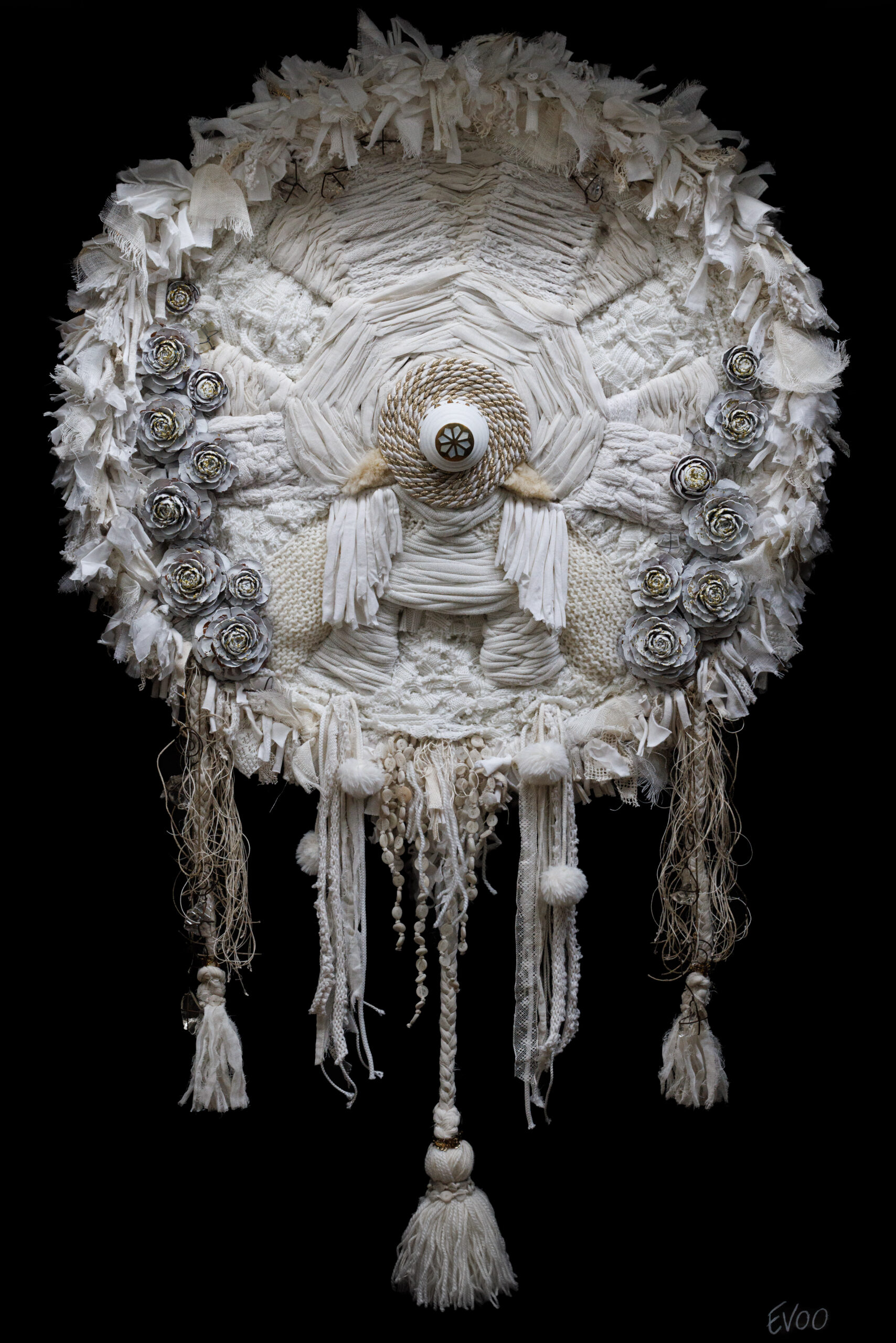 Ce chapeau blanc et complexe est orné de franges, de pompons et de roses en tissu, avec un motif central en forme d'œil.