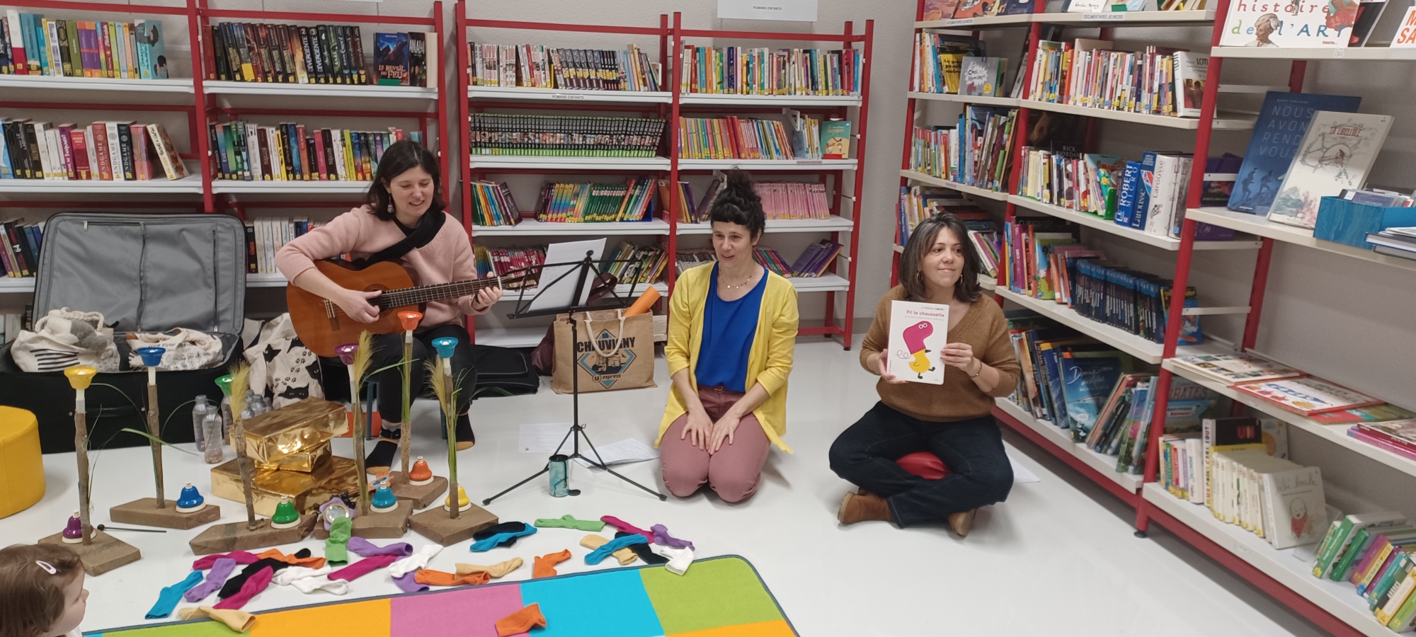 Trois femmes dans une bibliothèque. Une joue de la guitare, une autre tient un livre, et la troisième montre une illustrati
