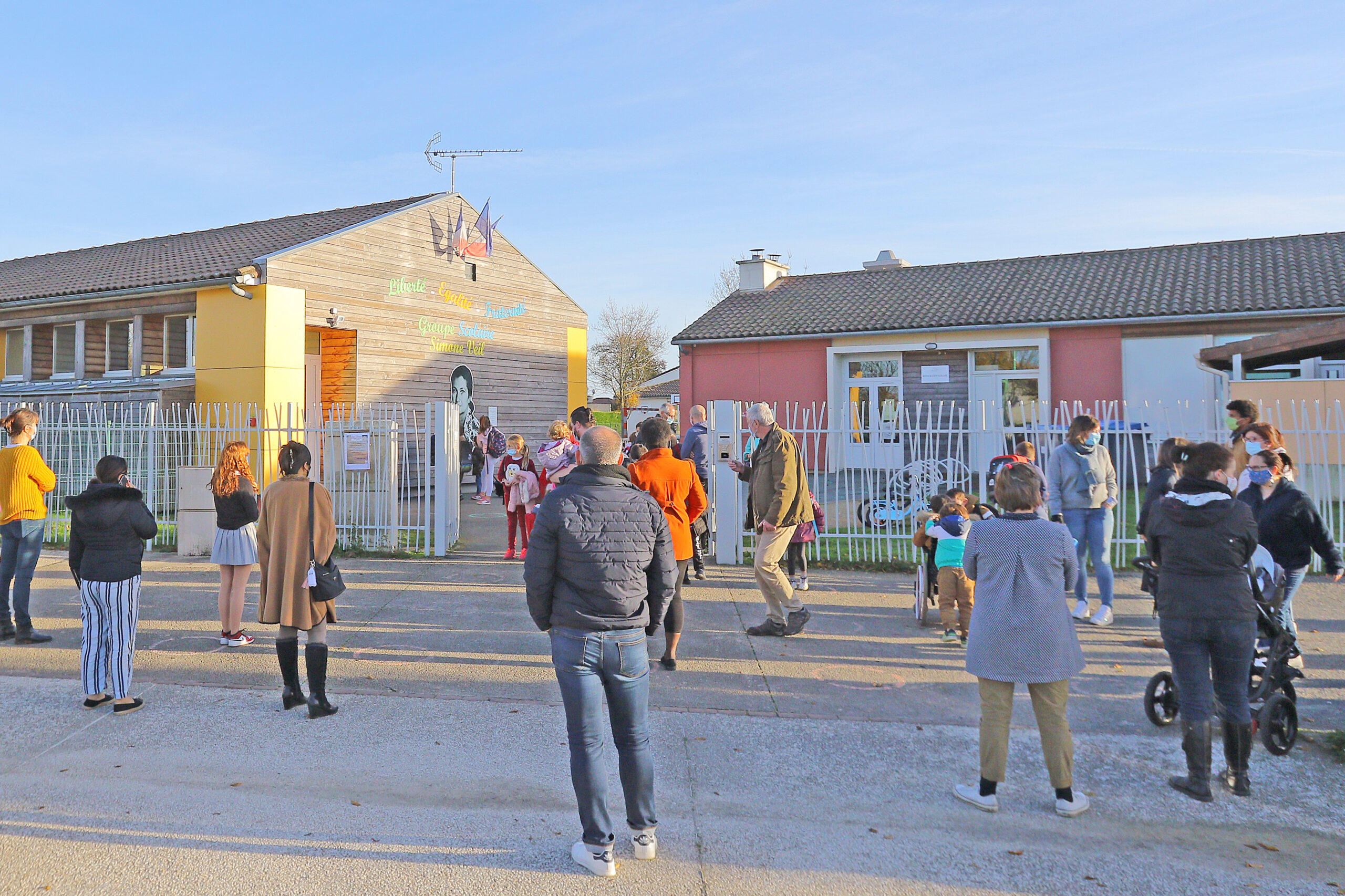 L'image montre un groupe de personnes, y compris des adultes et des enfants, se dirigeant vers une école colorée avec des b
