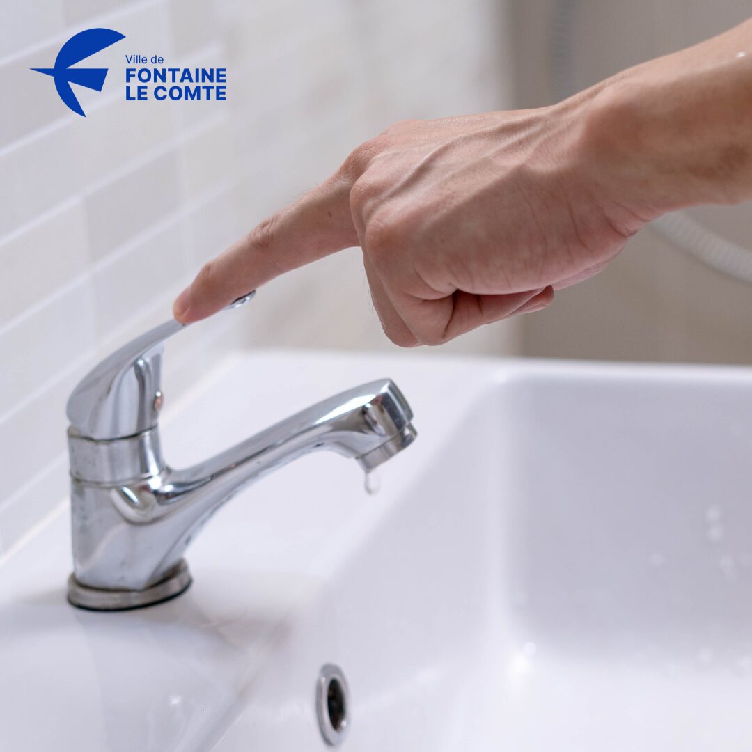 Une main tourne un robinet en argent sur un lavabo blanc.