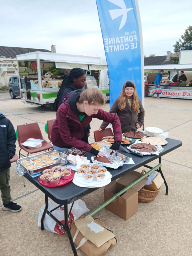 Trois personnes préparent des desserts sur une table à un stand de marché. Des gâteaux et des tartes sont exposés sur la