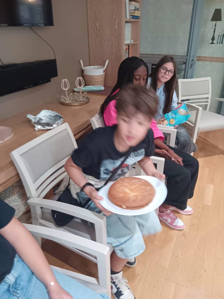 Trois enfants assis à table avec assiettes, découpant des aliments, dans une pièce lumineuse. Activité conviviale.