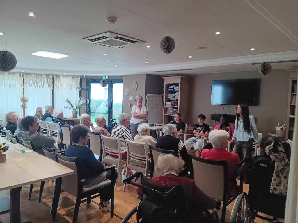 Groupe de personnes âgées participant à une réunion ou activité en salle, deux animatrices debout.