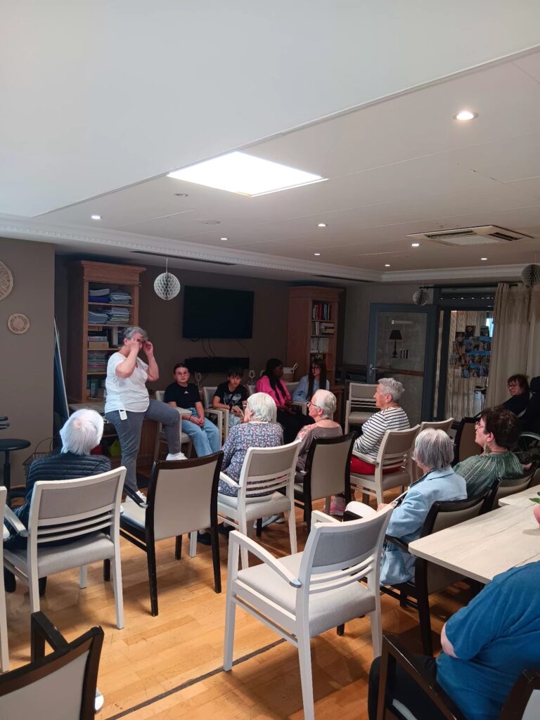 Groupe d’adultes assis en cercle écoutant une personne debout dans une salle conviviale, discussion ou atelier en cours.