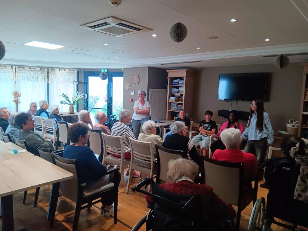 Groupe d’aînés assis en cercle dans une salle, écoutant deux personnes debout lors d’une réunion ou activité sociale