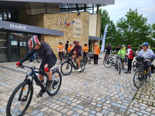 Groupe de cyclistes devant la mairie, prêts à partir pour une balade. Équipés et souriants, ils attendent sur une place p