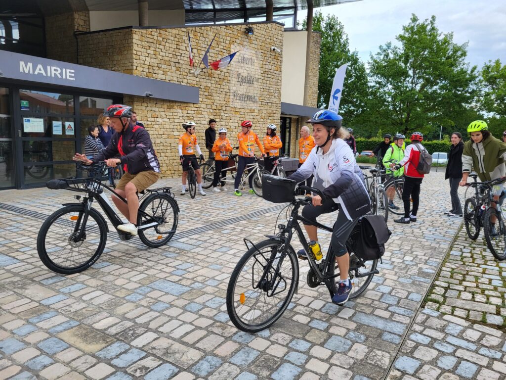Cyclistes devant la mairie, prêts à partir, équipés de casques, sur une place pavée.