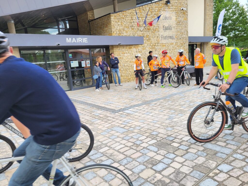 Cyclistes devant une mairie, groupe en tenue orange, accueil officiel, vélo, bâtiment en briques.