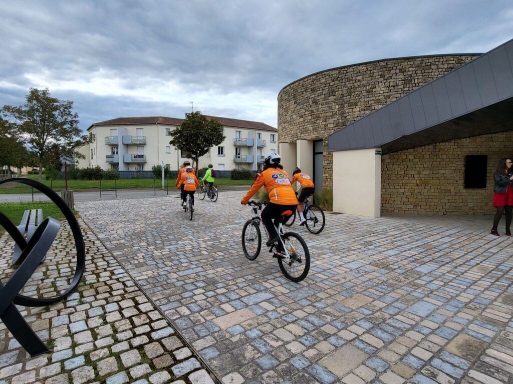 Cyclistes en tenues orange roulent sur une piste en pavés près d’un bâtiment moderne, sous un ciel nuageux.