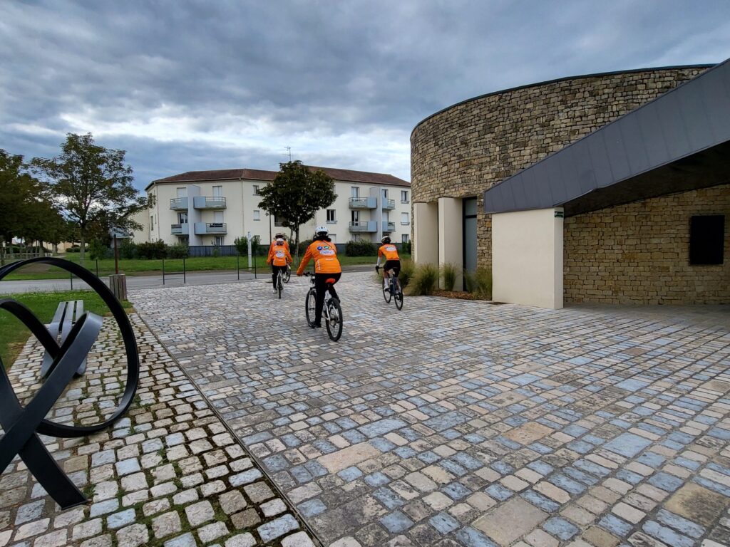 Trois cyclistes en tenues orange roulent sur une allée pavée près d’un bâtiment en briques sous un ciel nuageux.