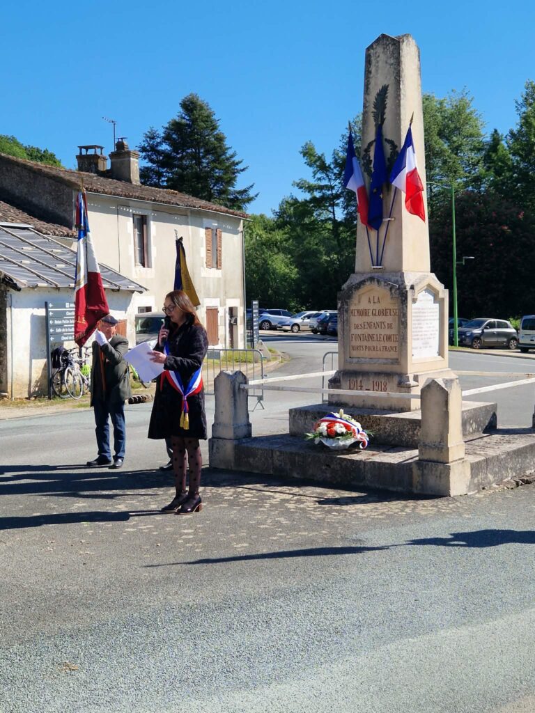 Cérémonie devant un monument aux morts 1914-1918, avec deux personnes, drapeaux français et fleurs, en hommage aux soldats