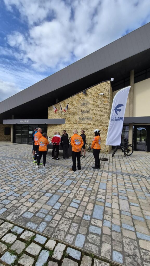 Un groupe de cyclistes en tenues orange devant la mairie de Fontaine-le-Comte, sous une bannière événementielle.