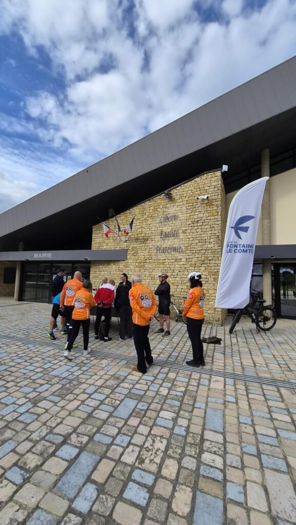 Groupe cyclistes en tenues orange devant mairie, bannière "Ville de Fontainebleau", ciel nuageux.