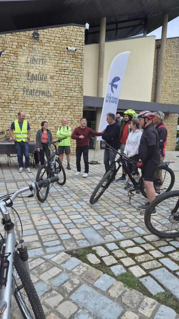 Groupe de cyclistes devant un bâtiment, banner "Fontaine le Comte", ambiance conviviale et départ d’un événement sporti