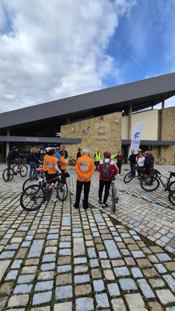 Groupe cyclistes devant mairie, vêtements colorés, ciel nuageux, pavés mouillés. Événement organisé.