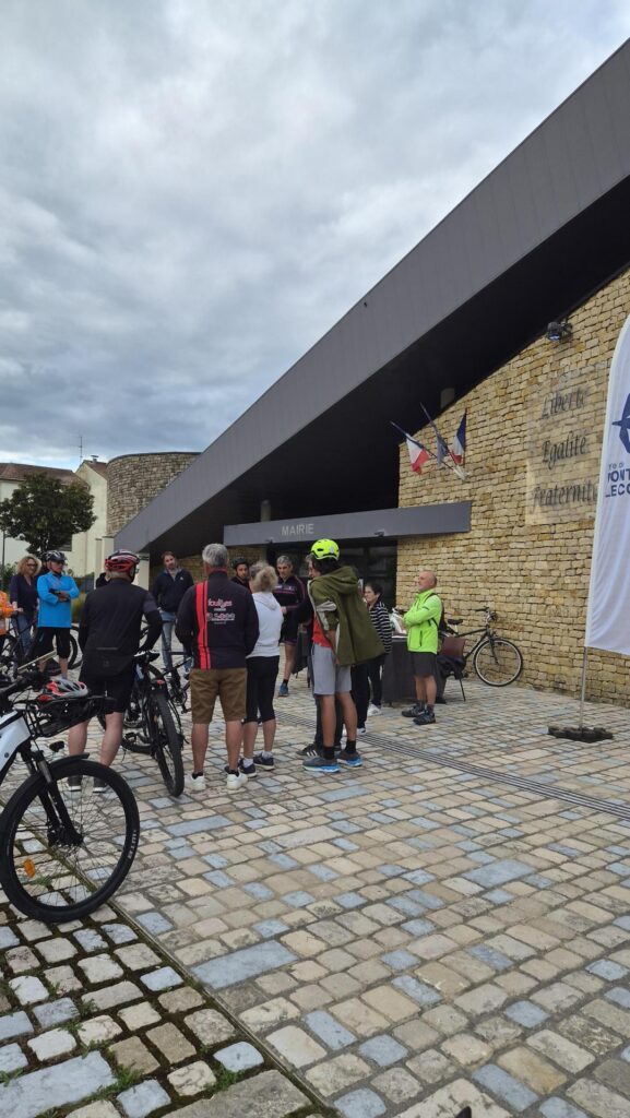 Groupe de cyclistes devant la mairie, sous un auvent, jour nuageux.