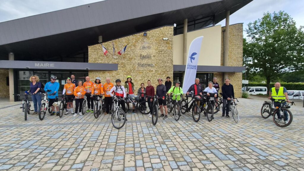 Groupe cyclistes posant devant mairie, avec drapeaux et bannières, prêt pour une sortie.