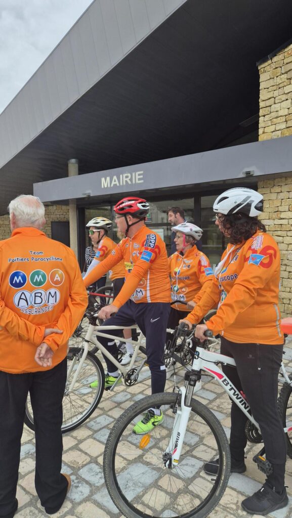 Cyclistes en tenue orange devant la mairie, discutant avec un organisateur, équipés de casques et vélos de course.