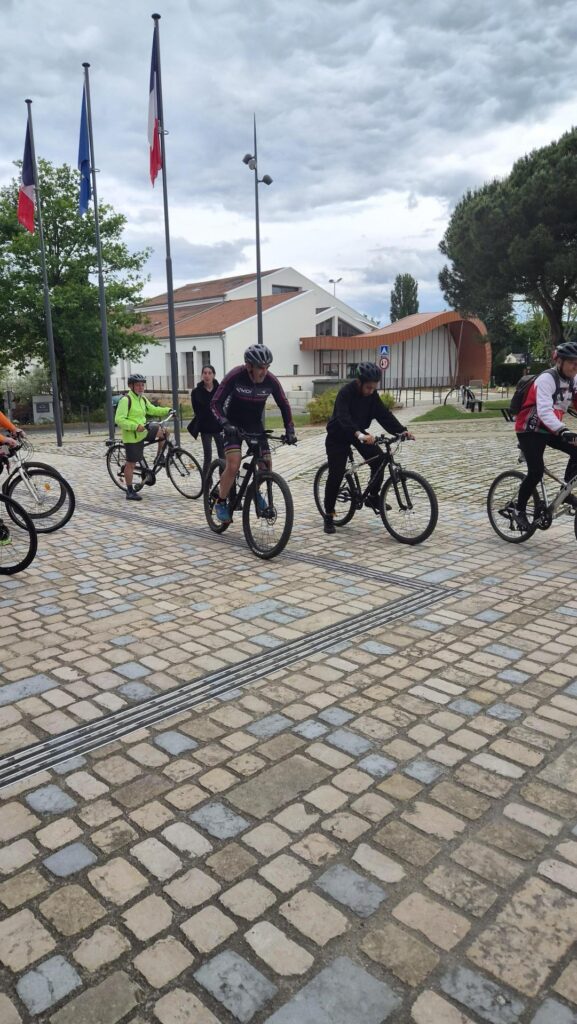 Cyclistes sur une place pavée avec bâtiments et drapeaux français en arrière-plan, temps nuageux.