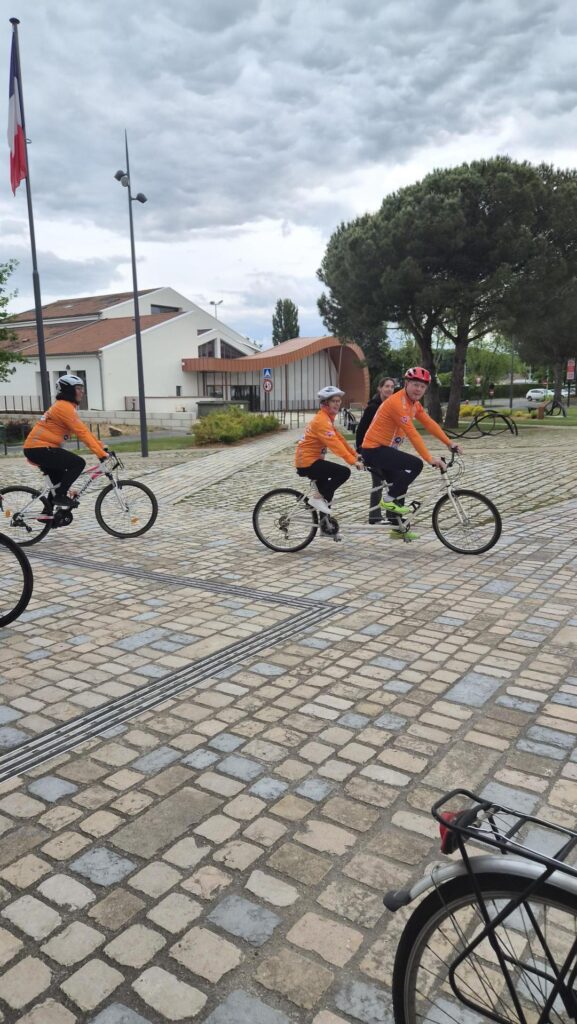 Trois cyclistes en tenues orange roulent sur une piste en pavés, sous un ciel nuageux, près d’un bâtiment et d’arbres.