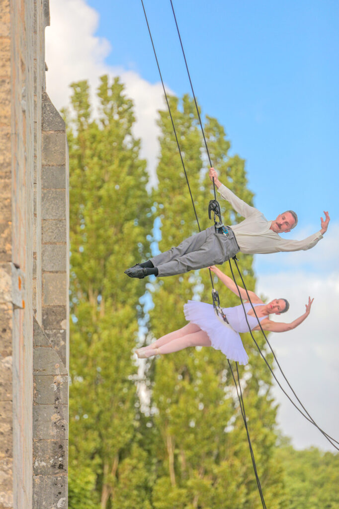 Deux artistes suspendus en équilibre sur fil, l’un en costume, l’autre en tutu, devant un arbre et un mur.