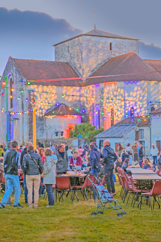 Fête en plein air devant un vieux bâtiment illuminé, tables et chaises, groupe de personnes en conversation.