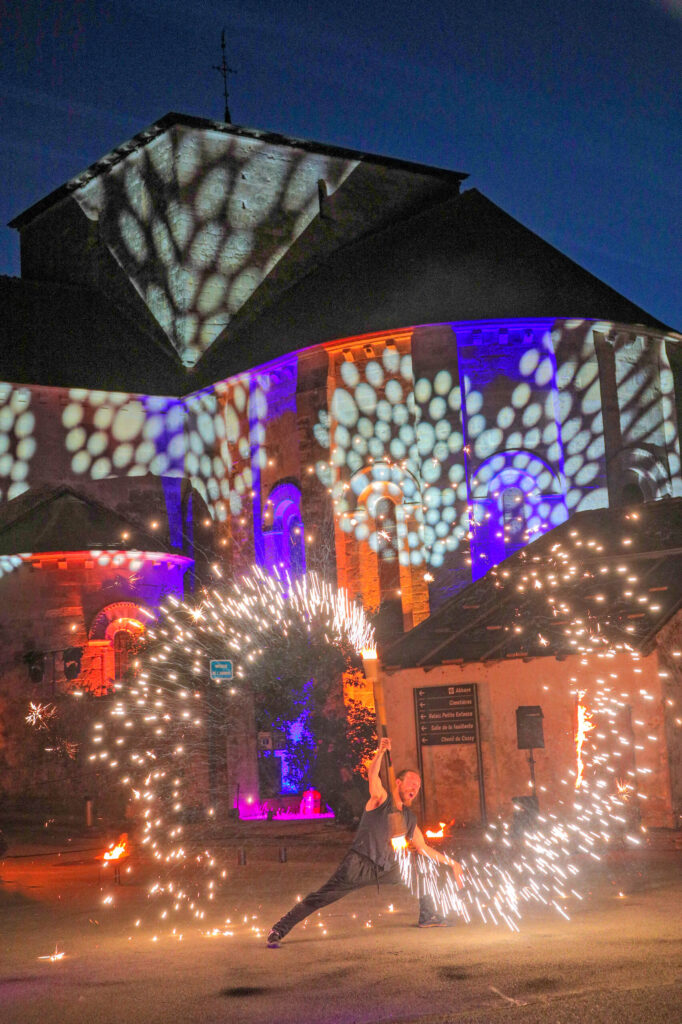 Artiste manipulant des torches lumineuses devant un bâtiment illuminé de motifs colorés, nuit étoilée.