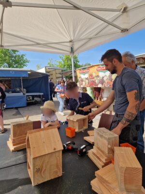 Des enfants et adultes assemblent des nichoirs en bois sous une tente lors d’un événement extérieur.