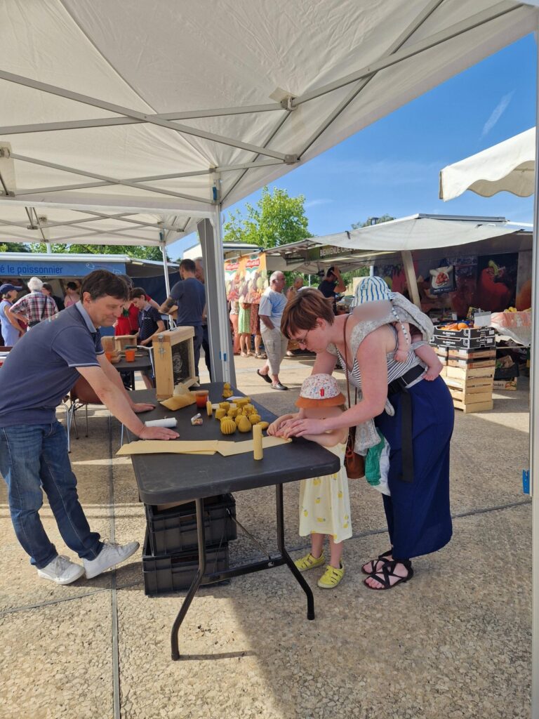 Un homme aide une enfant à fabriquer des petits pains sous une tente de marché, avec une femme et des produits artisanaux a