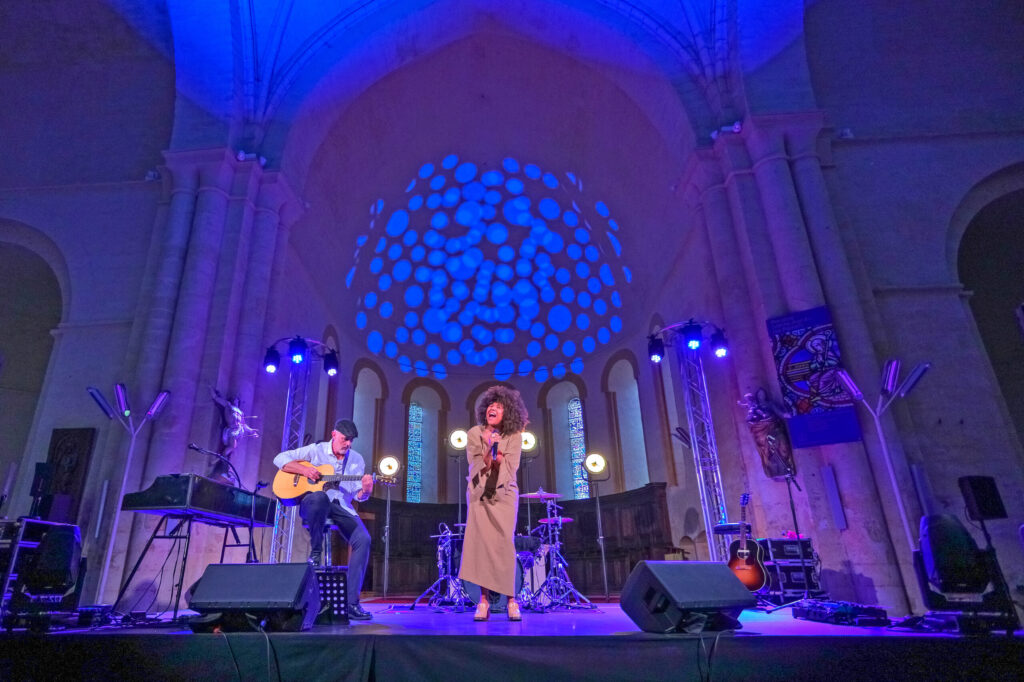 Concert en intérieur : musiciens sur scène éclairée, guitariste et chanteuse devant un public, ambiance lumineuse bleuté