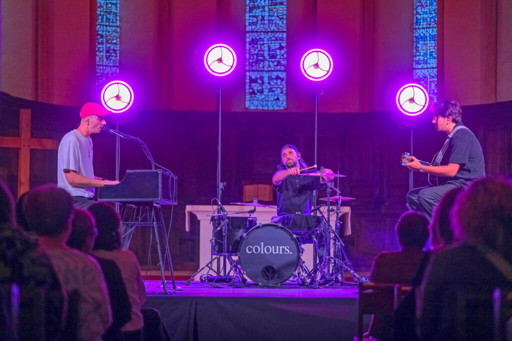 Trois musiciens jouent sur scène sous une lumière violette, avec un claviériste, un batteur et un guitariste devant un pub