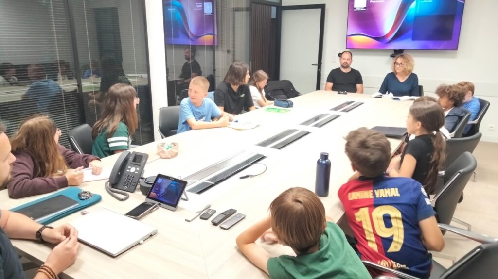 Réunion en salle avec enfants et adultes autour d’une table, discutant avec écrans et documents.