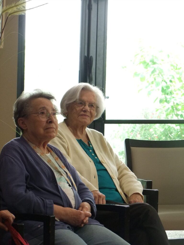 Deux personnes âgées souriantes assises près d'une fenêtre, vêtues de cardigans, en intérieur lumineux.