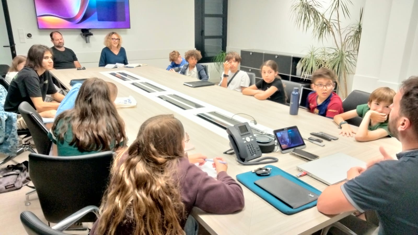 Réunion en salle avec enfants et adultes assis autour d’une table, discutant avec écrans et documents.