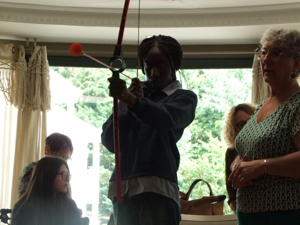 Une personne lance une balle avec une corde dans une pièce, observée par un groupe attentif près d’une fenêtre.