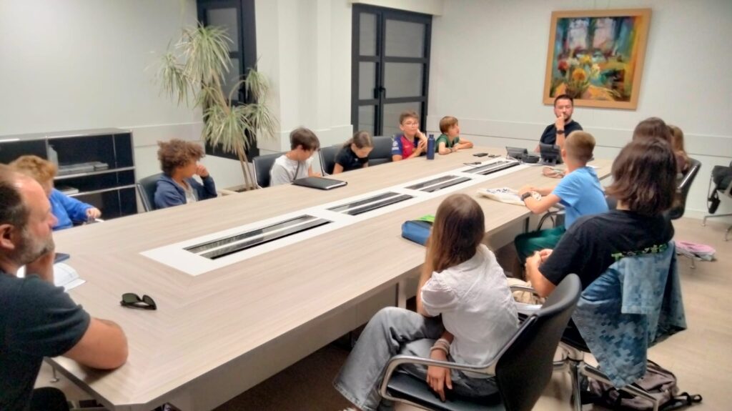 Groupe en réunion assis autour d’une grande table, discutant dans une salle moderne avec plantes et tableau décoratif.