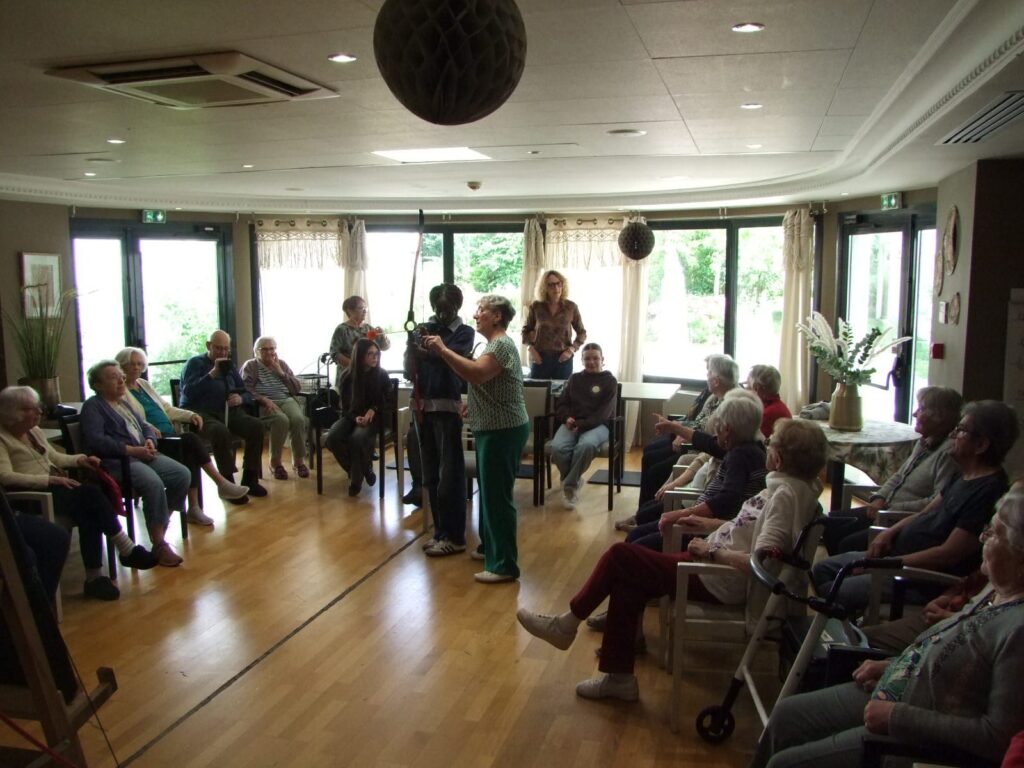 Groupe d’adultes assis en cercle dans une salle lumineuse, écoutant une personne debout avec un instrument de musique.