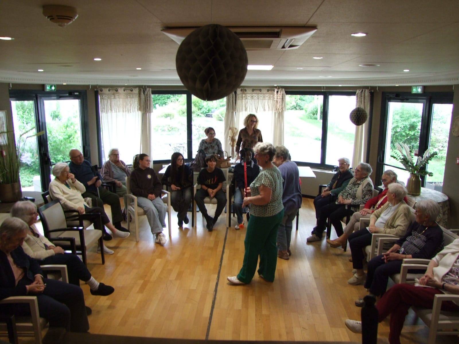 Groupe de personnes assises en cercle dans une salle lumineuse, écoutant une animatrice debout au centre.