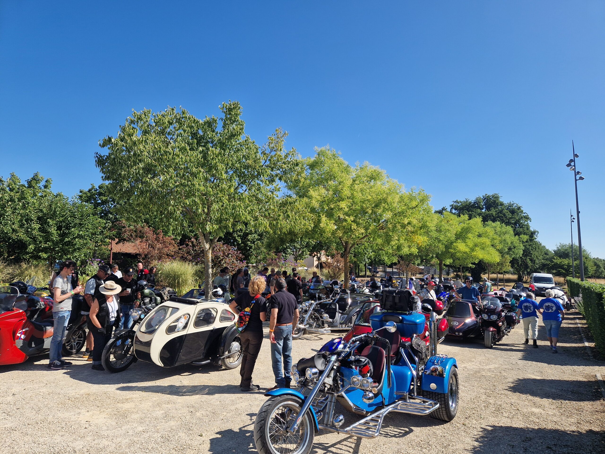 Groupe de motards et triporteurs réunis lors d’un rassemblement en plein air sous un ciel dégagé, entourés d’arbres.