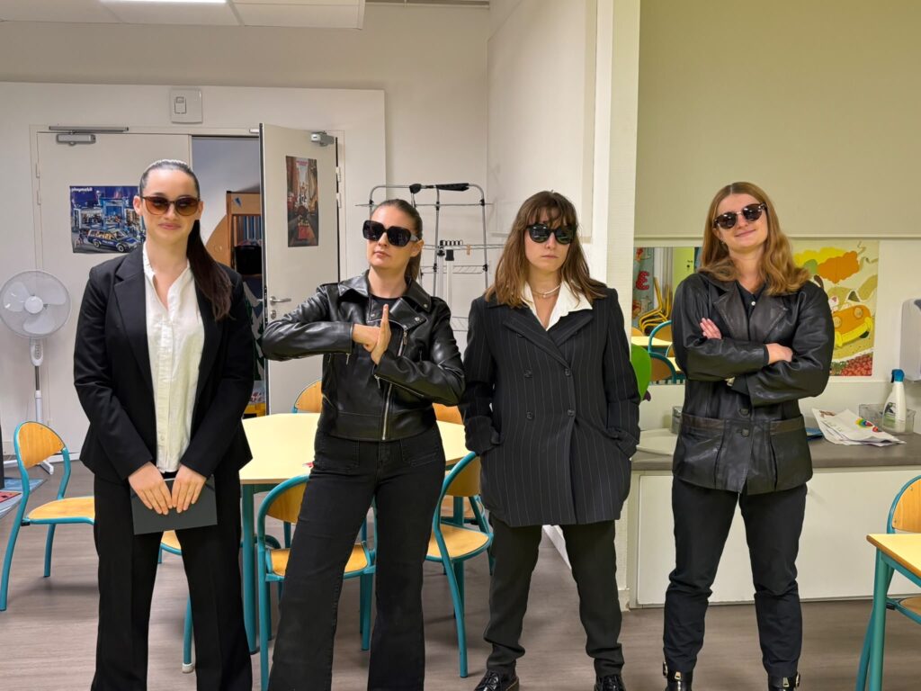 Quatre femmes posant en intérieur, vêtues de tenues noires, lunettes et poses confidentes, salle avec tables et chaises.