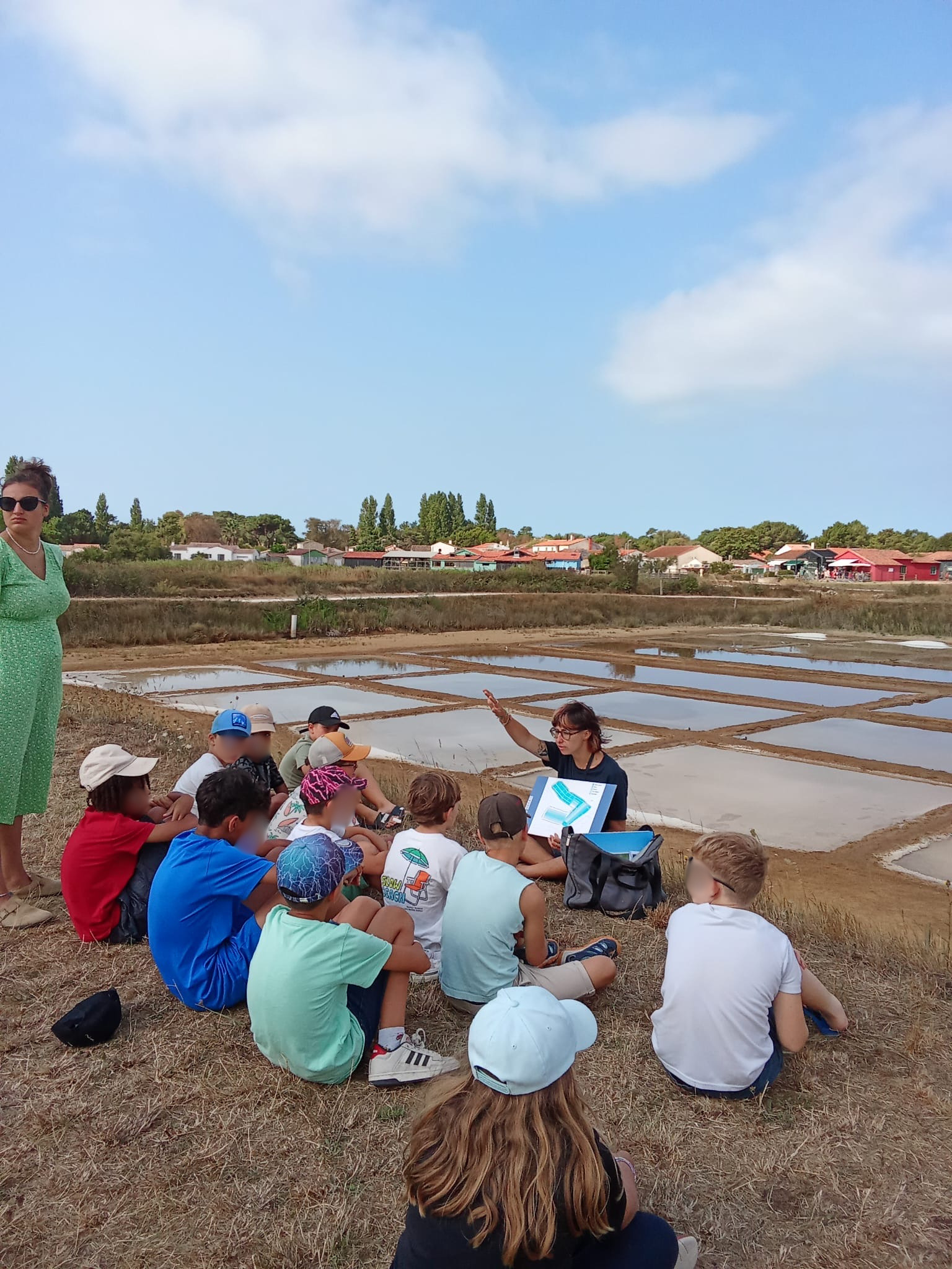 Un groupe d’enfants assis écoute une guide près de bassins de sel, sous un ciel dégagé. Activité éducative en extéri