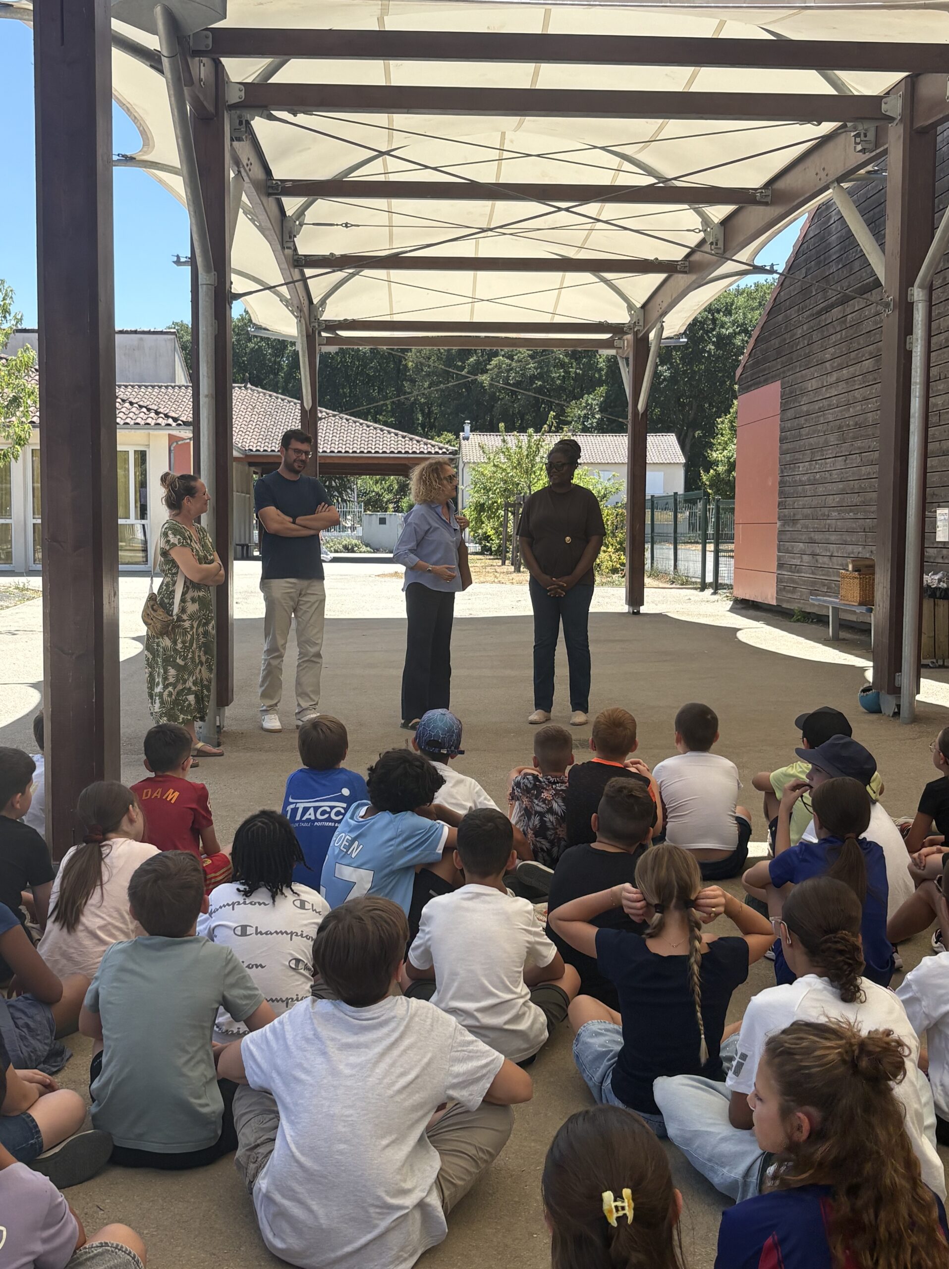 Groupe d'enfants assis écoutant quatre adultes sous une pergola en extérieur, scène éducative ou réunion scolaire.