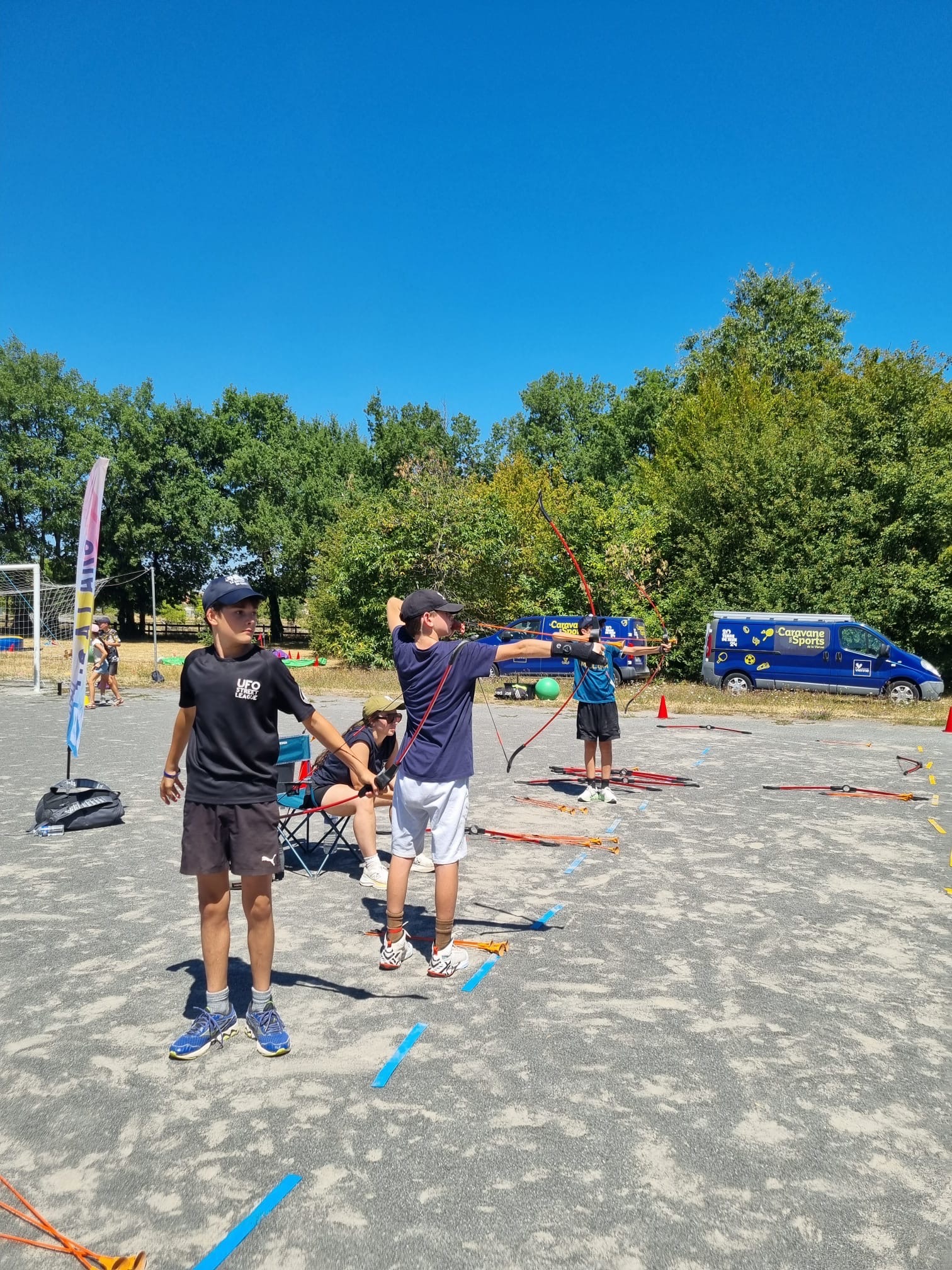 Enfants apprenant à tirer à l’arc en extérieur sous un ciel bleu, avec du matériel et des cibles au sol.