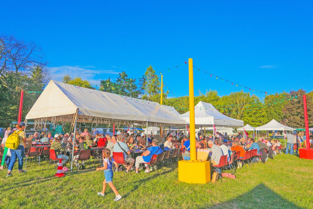 Fête en plein air sous tentes avec nombreux convives assis, enfant en mouvement, décorations lumineuses, ambiance joyeuse.