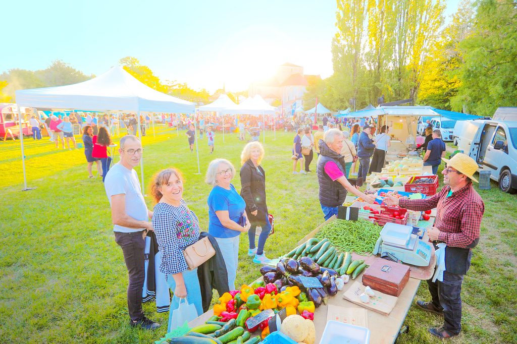 Femmes et hommes souriants à un marché en plein air, entourés de légumes frais et de stands sous tentes.
