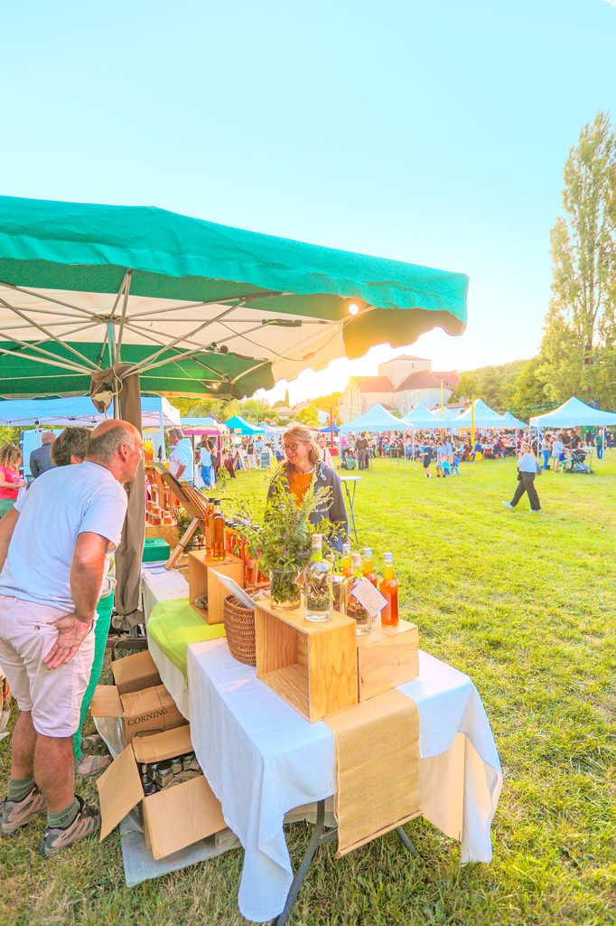 Un stand de marché sous tente verte avec des produits artisanaux, une vendeuse souriante et des clients en plein air.