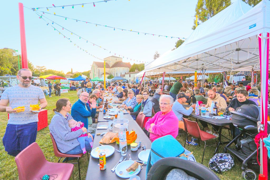 Groupe de personnes partageant un repas convivial sous une tente festive, avec décorations lumineuses et vue sur un village.