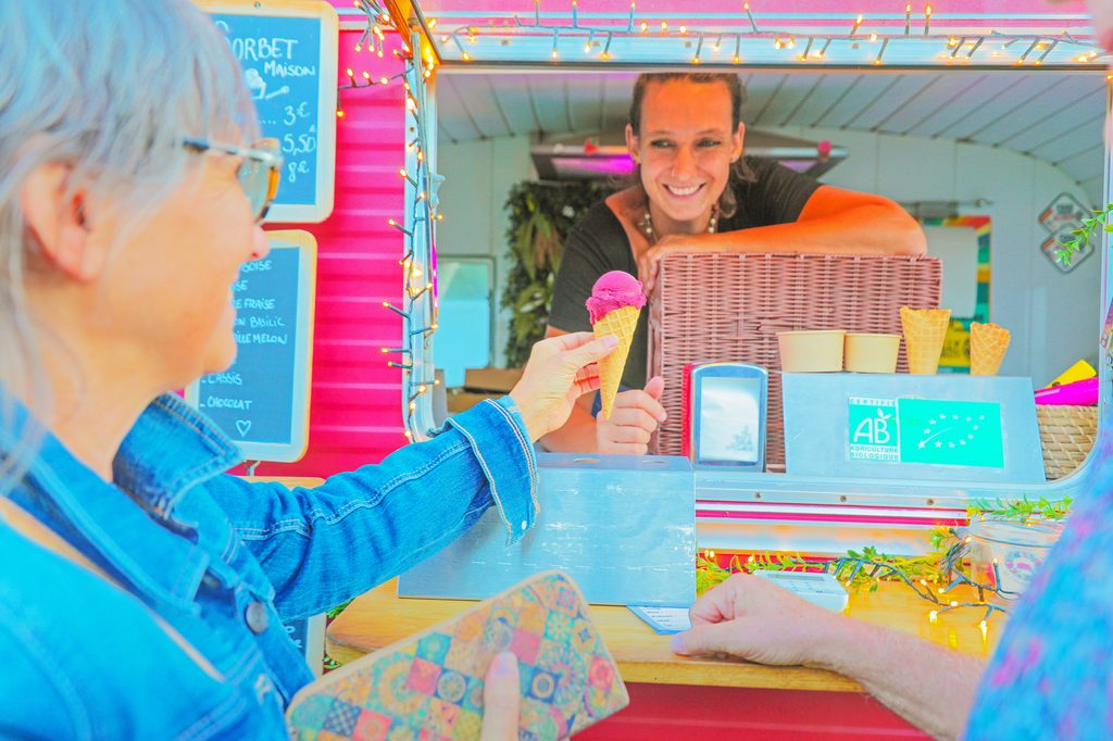 Une personne reçoit un cornet de glace colorée d’une vendeuse souriante dans un stand gourmand, décoré de guirlandes lu