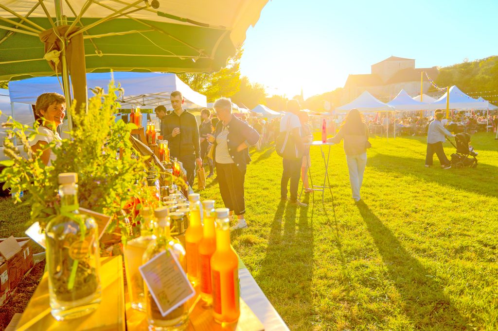 Fête en plein air avec dégustation de vins sous tente, ensoleillée, participants souriants et bouteilles exposées.