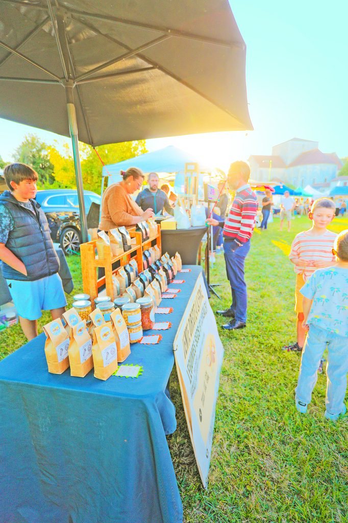 Stand de vente sous tente avec produits artisanaux, enfants et vendeurs en extérieur ensoleillé.